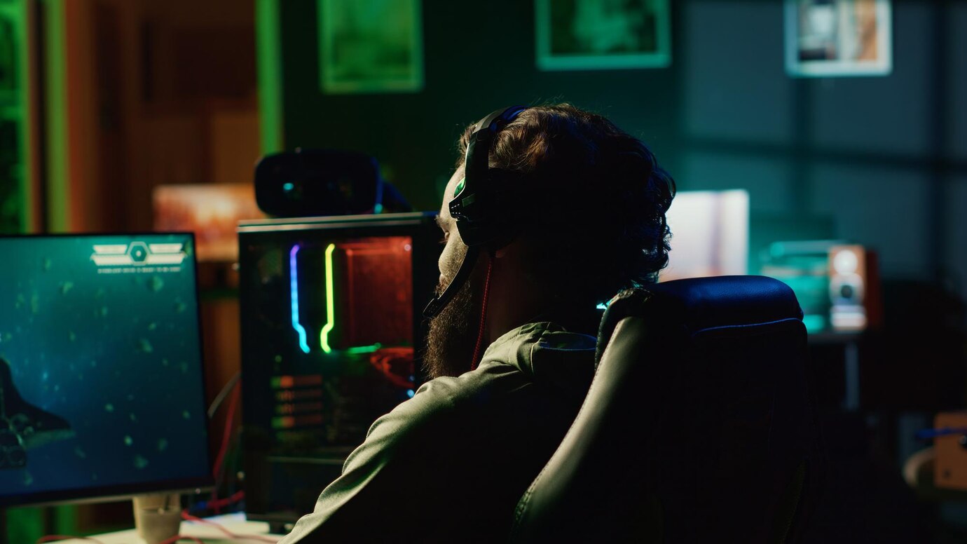 A person wearing headphones sits at a desk, facing a computer with colorful LED lights, playing a game