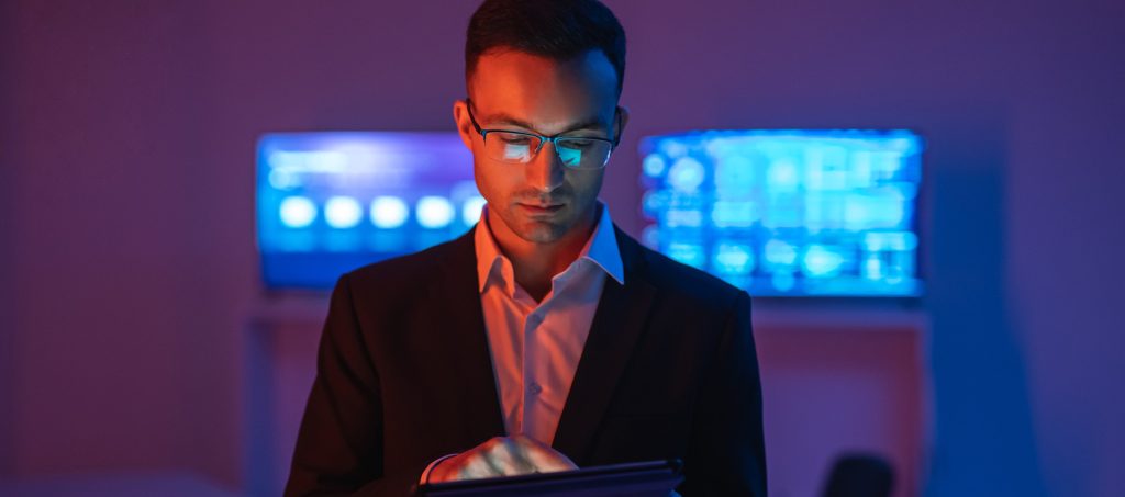 Man in a suit using a tablet in a dimly lit room with blue-lit monitors in the background.