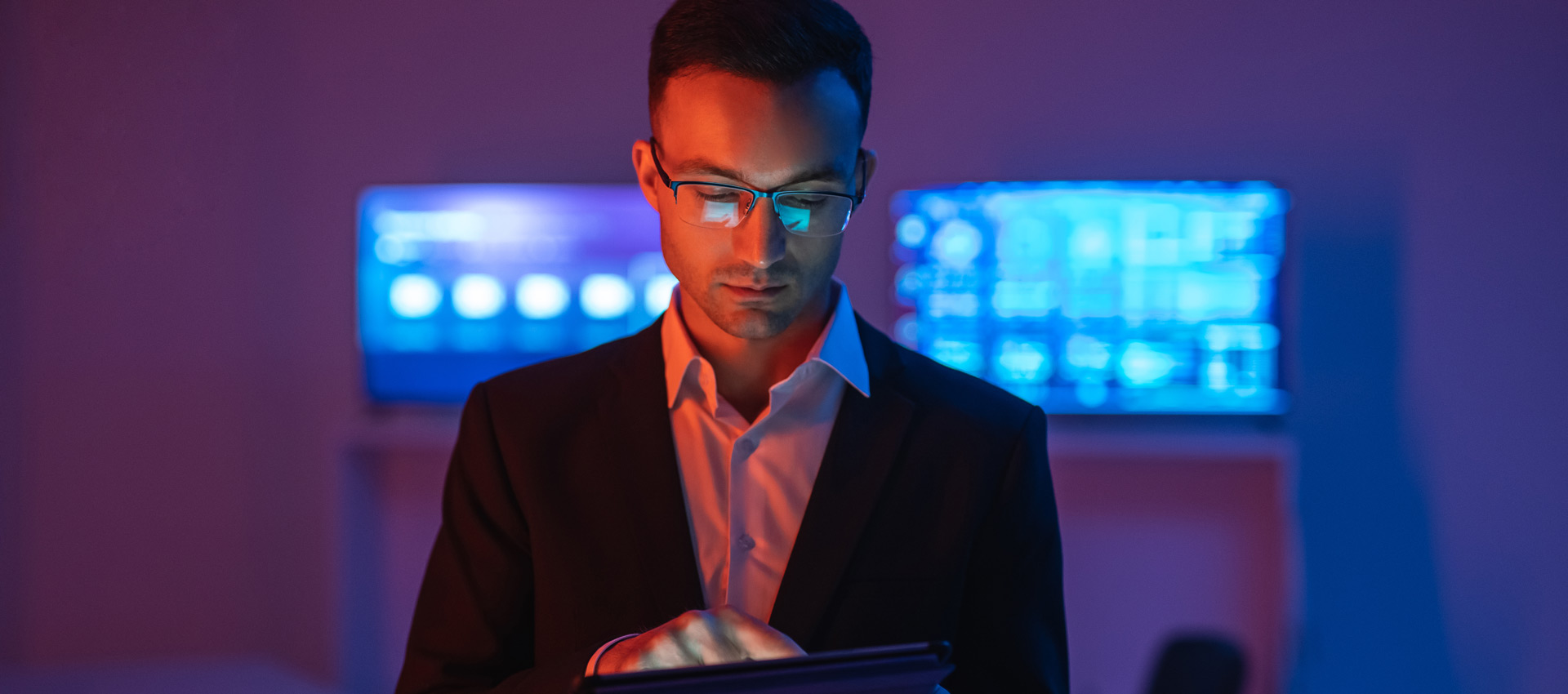 Man in a suit using a tablet in a dimly lit room with blue-lit monitors in the background.