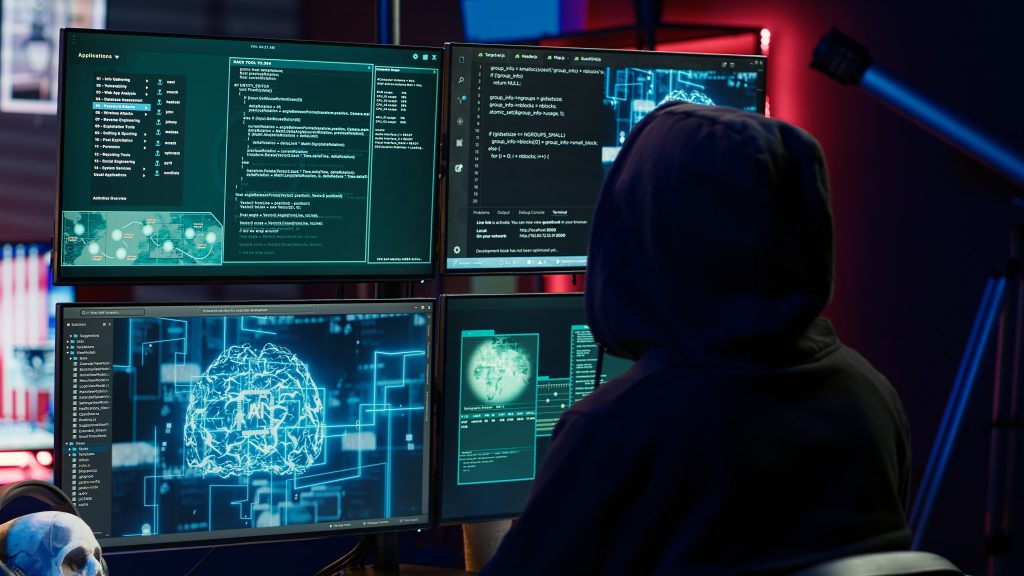 A person in a hoodie monitors multiple computer screens, deeply focused on cybersecurity operations.